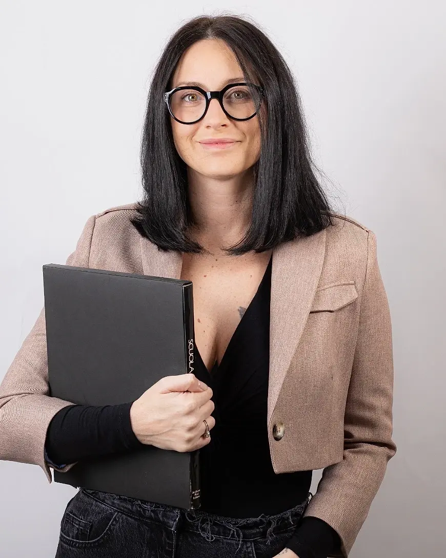 Professionista con occhiali tondi, giacca rosa e cartellina nera su fondo chiaro, ritratto di Ilia Da Lozzo